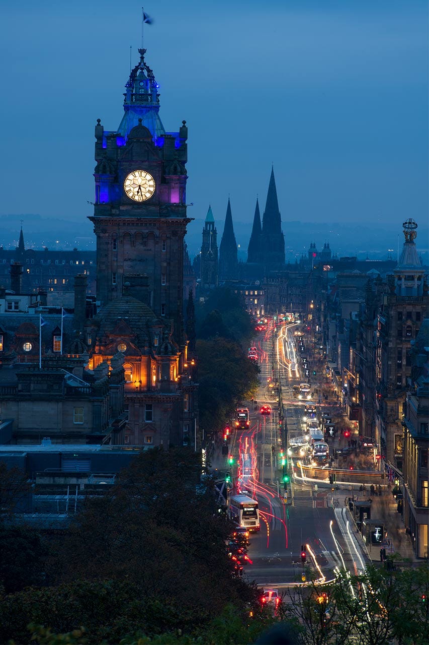 A big purple squarish clock tower on the left and a busy street with vehicle lights stretching away on the right against a blue dusky sky