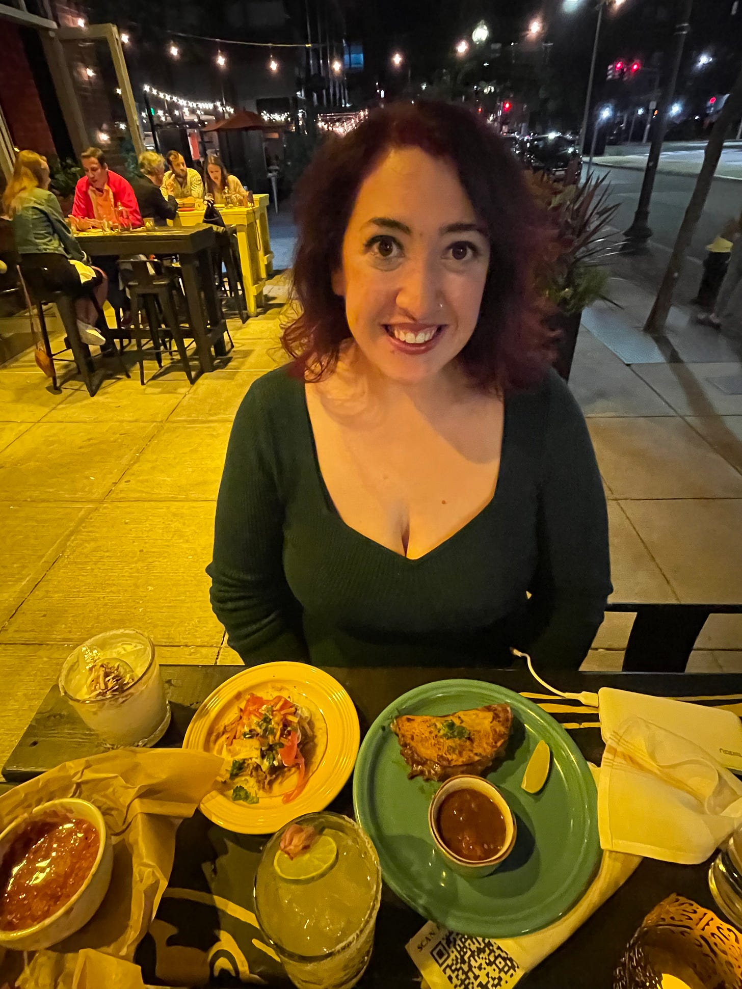 A woman sits outside at night at a table with a plate of tacos in front of her. She has short red hair and a nose ring. 