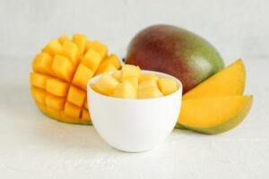 Small bowl with mango pieces and cut mangoes on white background, closeup