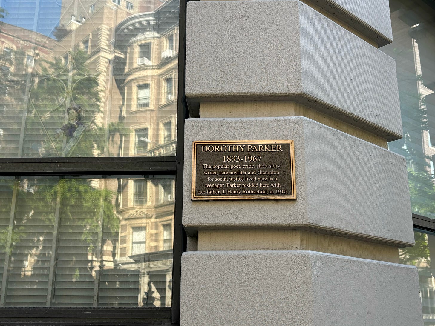 A close up of the black on the West 80th Street building. It is on the wall next to a window. There is a doll propped up against the protective frame in the window and the buildings across the street are reflected. The plaque reads: "DOROTHY PARKER 1893-1967 The popular poet, critic, short story writer, screenwriter and champion for social justice lived here as a teenager. Parker resided here with her father, J. Henry Rothschild in 1910."