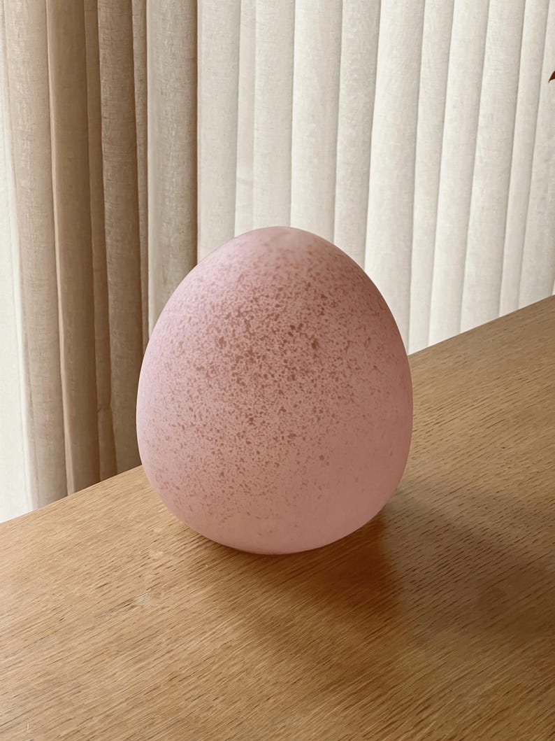 May include: Close-up view of a speckled, light pink egg-shaped decorative object. The object has a slightly rough, textured surface and appears to be made of glass or a similar material. It is sitting on a light brown wooden surface.