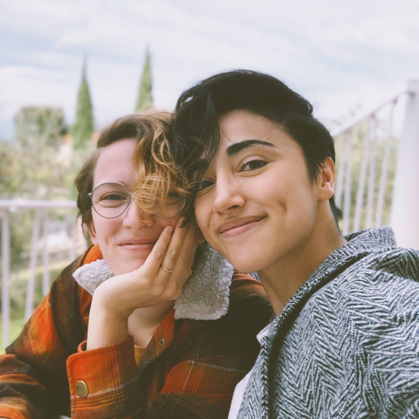 Torri and Alex smiling at the camera in a selfie. Their heads are close together and they look content. Behind them are some trees and a cloudy sky.