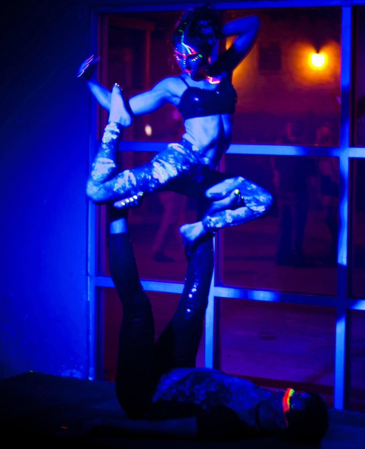 Lyric doing AcroYoga, they are balanced on the feet of a tall base, doing a complex pose. They scene is a rave style party, with black lights and glow paint. 