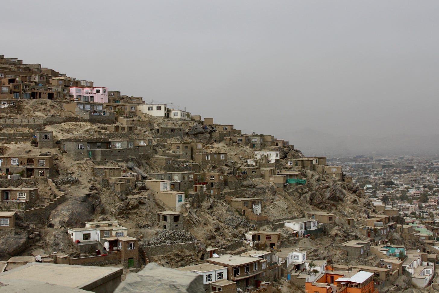 Brown mountain peak rises in Kabul with rudimentary houses clinging to the side of the peak and dense development on the valley below