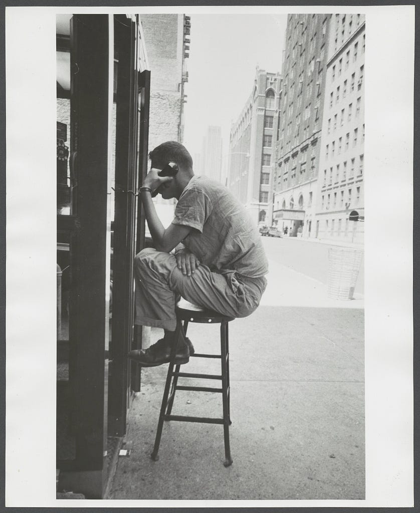 Photo shows: Man, full-length portrait, seated on stool outside a telephone booth, phone raised to ear, view of buildings in background.