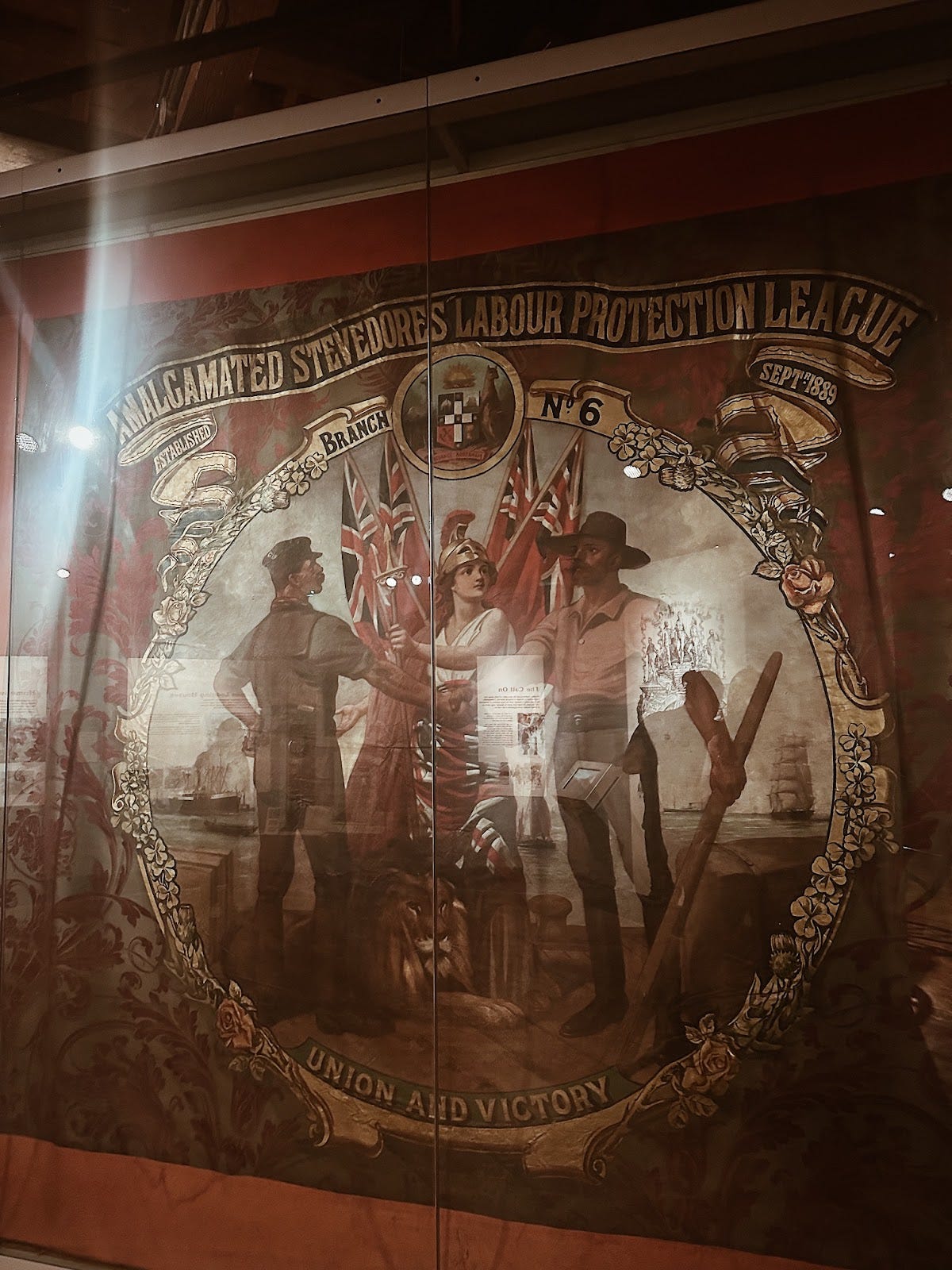 a photo of a tall and wide flag in a case. the brown and red flag depicts 2 figures shaking hands and a symbolic figure between them in front of australian and british flags below a banner reading "amalgamated stevedores labour protection league"