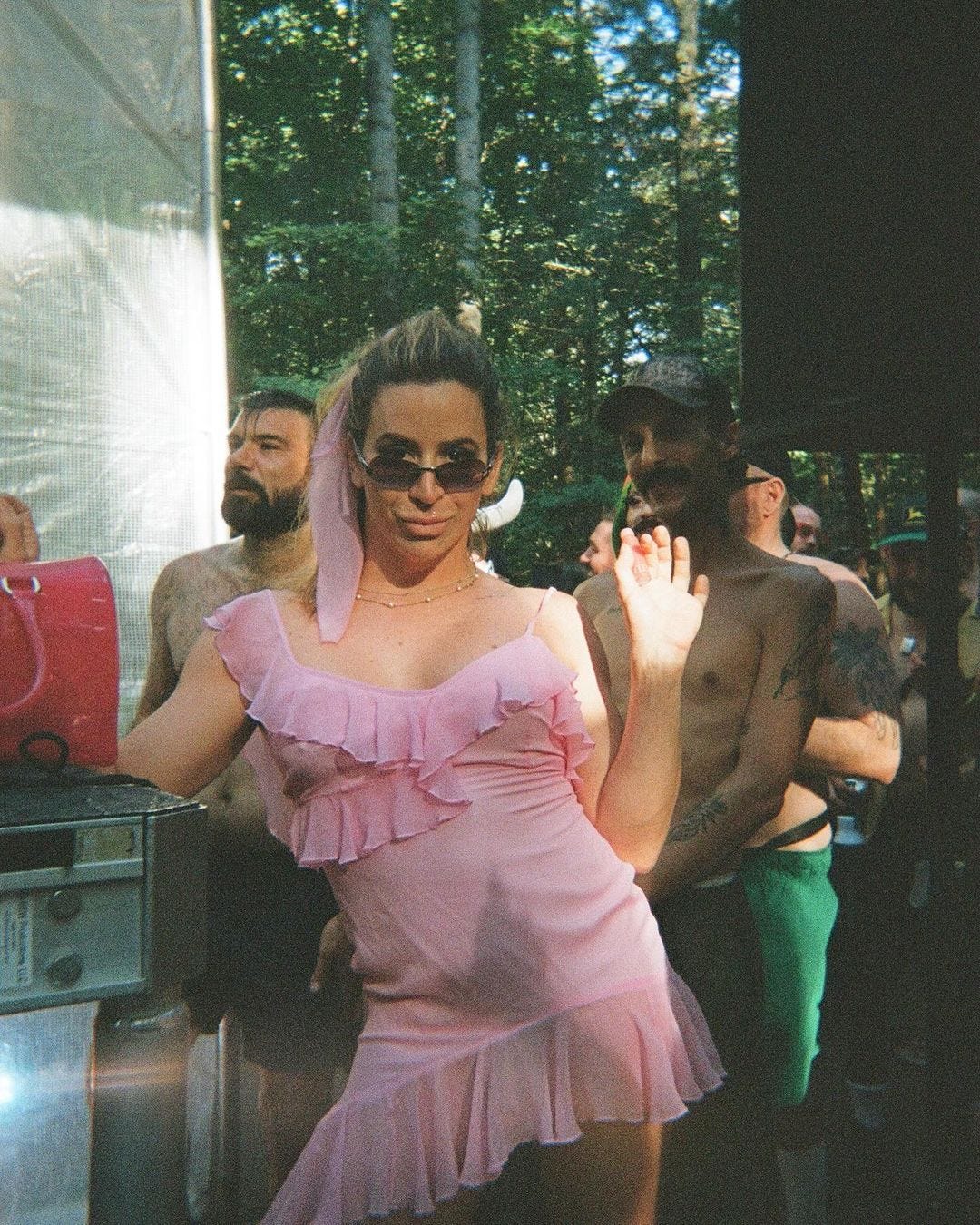 A disposable camera photo of a white trans woman dappled in afternoon sunlight. She is wearing a pink dress with ruffle detailing. The same diaphanous fabric is tied into her pony. She has 2 thin gold necklaces. One of her hands is on a translucent pink handbag. The other is holding a stick of pink chapstick she is about to apply to her pouting lips. Her weight is on one hip, popped outShe wears sunglasses low on her nose, below her eyes. Her face, locked into the camera, looks like a kind of girlfriend who would sleep with her boyfriend’s fine ass best friends while he’s away in the army.