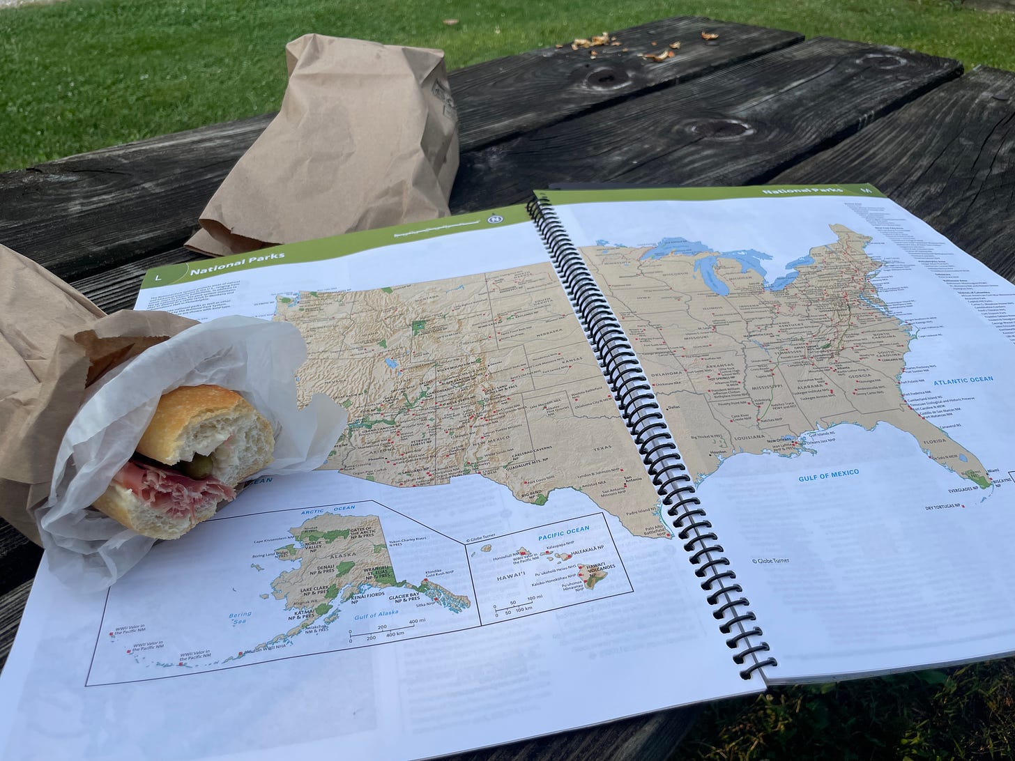 Photograph of a map of the United States laid out on a picnic table, next to a sandwich in a brown paper bag.