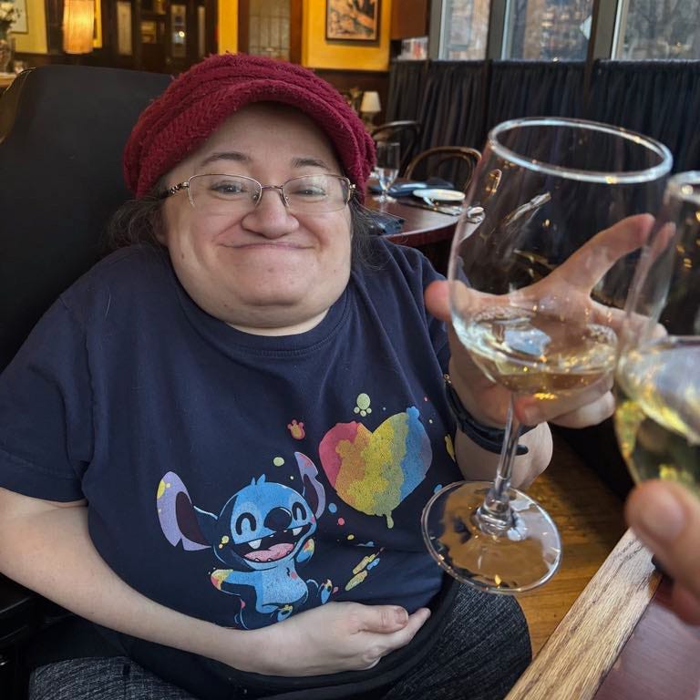 Athena in a restaurant raising her white wine glass in a toast. He's wearing her red knit cap with a brim and a t-shirt with Disney's Stitch painting a rainbow heart.