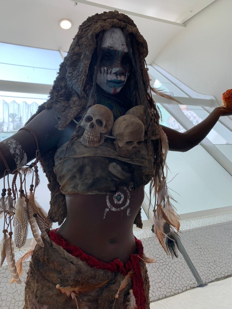 A woman dressed in animal skins, feathers, and a necklace of human skulls. She holds fire in her bare hands