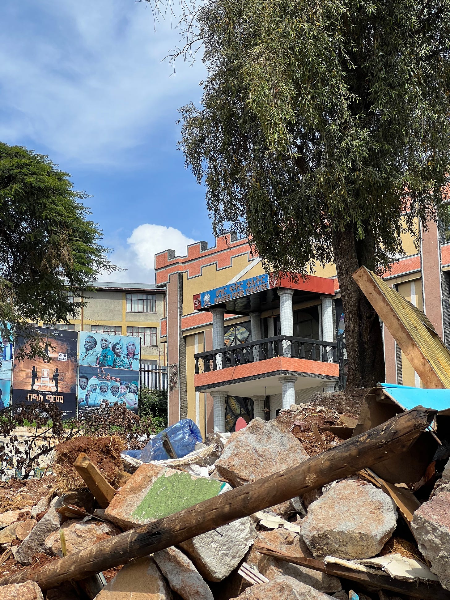 A colorful building with one big balcony has a sign on it that reads Hager Fikir Theater in English and in Amharic. There are theater poster of three shows on the right side and a tree on the right side. The view is half covered by rubble from the demolition of the gates of the theater house. 