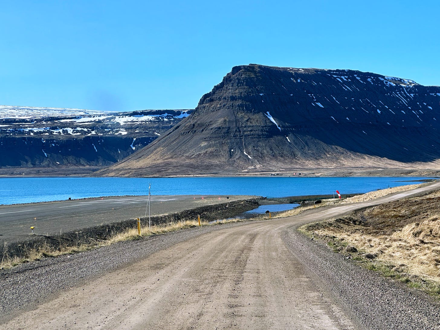 A gravel road curving around bright blue water with snow capped mountains in front and a cloudless blue sky overhead.