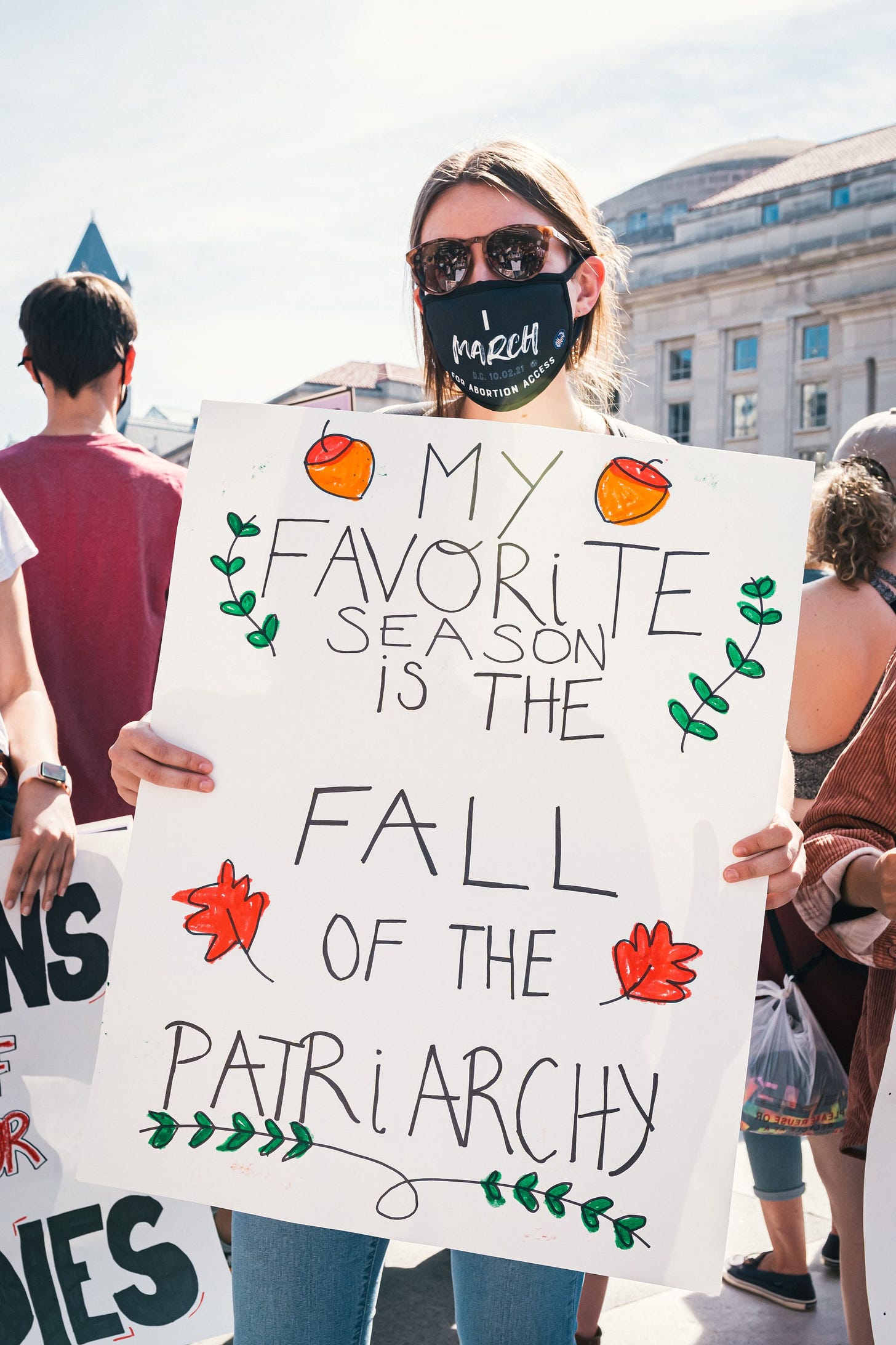 A woman stands at a protest, wearing a mask and holding up a sign that reads: my favorite season is the fall of the patriarchy