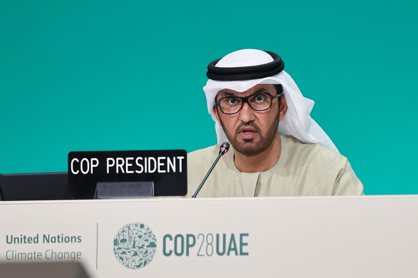 In a photo, a Saudi man wearing glasses and stubble sits at a desk in front of which is a sign that reads "COP president".