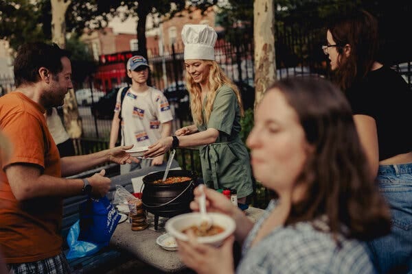 A woman with long blond hair wearing a white chef’s hat serves stew from a black cauldron in a New York City Park.