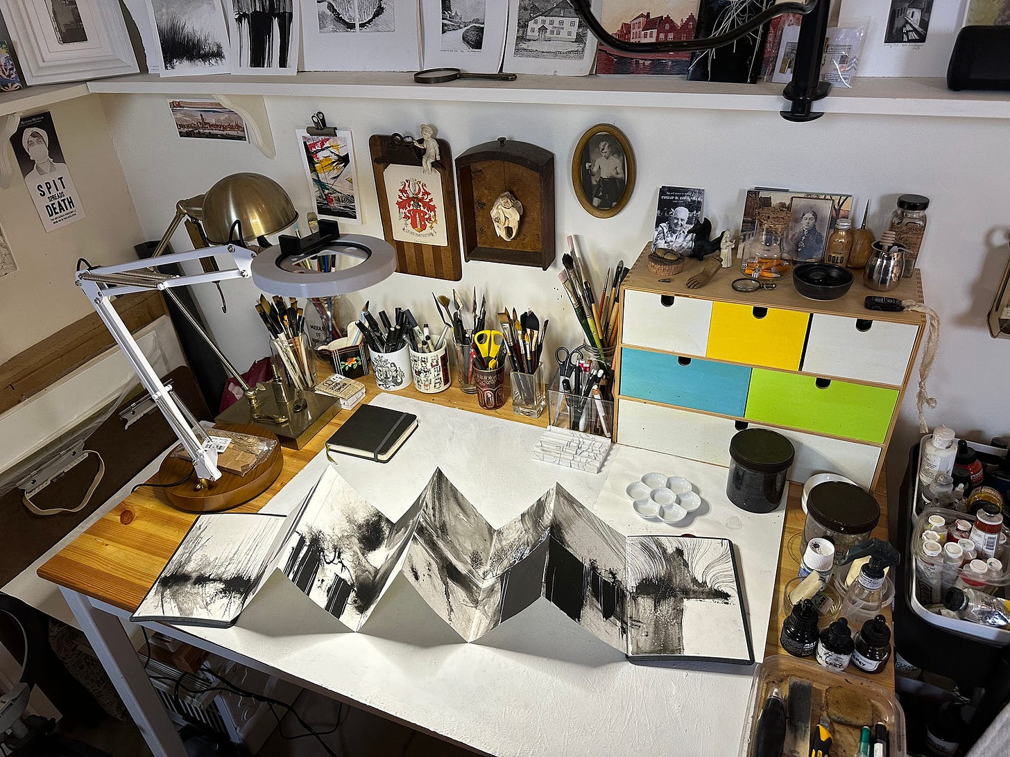 Photo of my desk, taken from a high angle. A semi- open accordian-shaped sketchbook is lying across the desk, showing black and white abstract black and white ink paintings. Loads of art supplies are around the edges of the desk, including many jars holding pens, pencils, brushes. A ring light sits to the left side of the desk, and lots of art rests on a slender shelf above the desk.