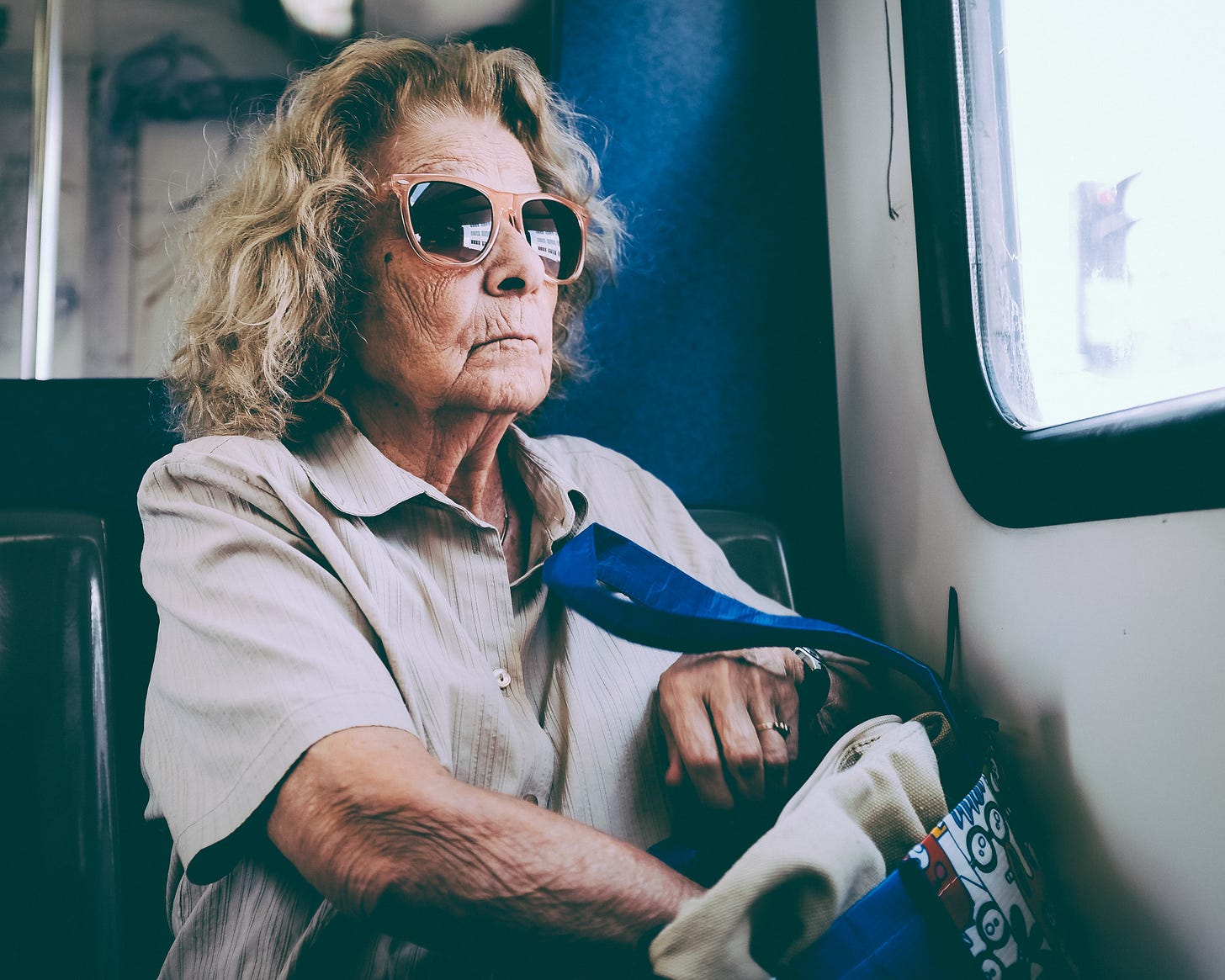 Imagem: retrato mulher idosa, cabelos loiros, óculos escuros olhando na janela de um ônibus