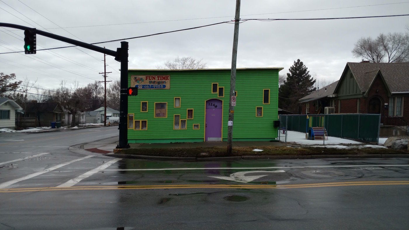 Photo of a small green building on the corner of an intersection. Daytime, cloudy sky. It looks like an average small town, the other visible buildings are houses. The daycare is small, with green paneling and lots of randomly placed little rectangular windows. It has a purple door.