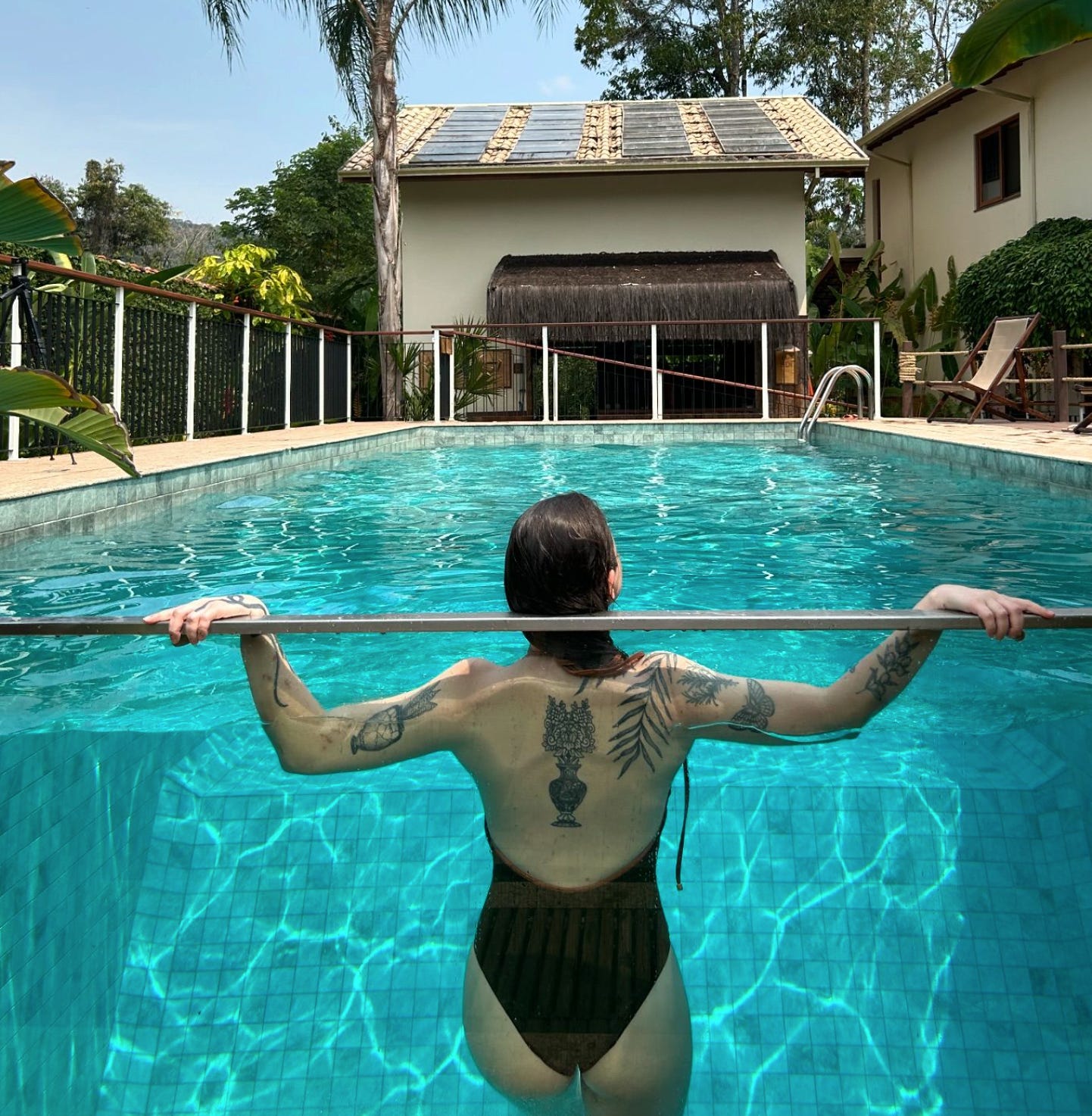 Euzinha, toda branquela, porém tatuada, de costas em uma piscina cuja lateral é feita de vidro, dando para ver debaixo d'água, trajando um maiô verde que deixa as costas aparecendo. 