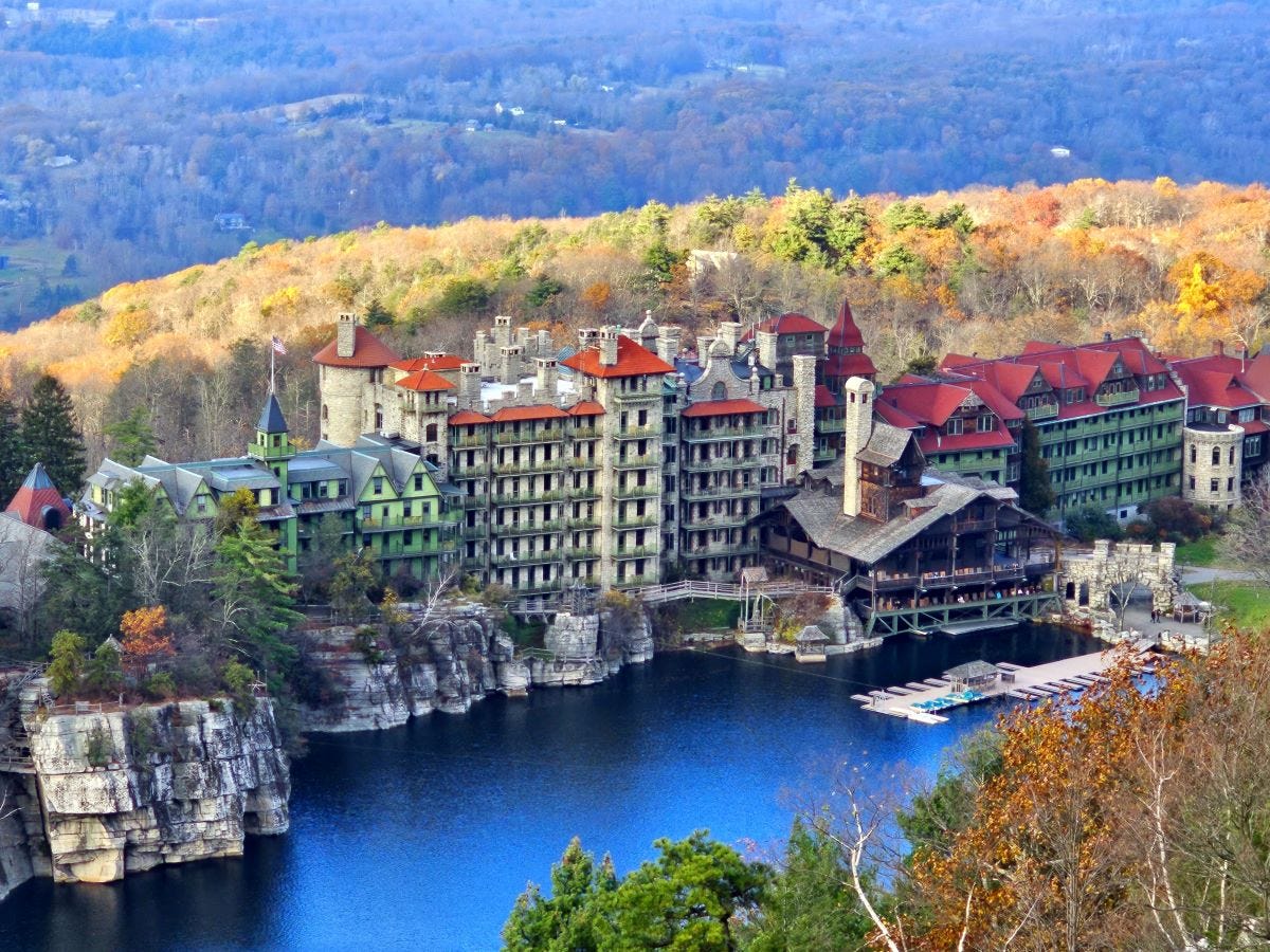 The Mohonk Mountain House a sprawling castle-like structure, with mountains in the background an a blue lake in the foreground.