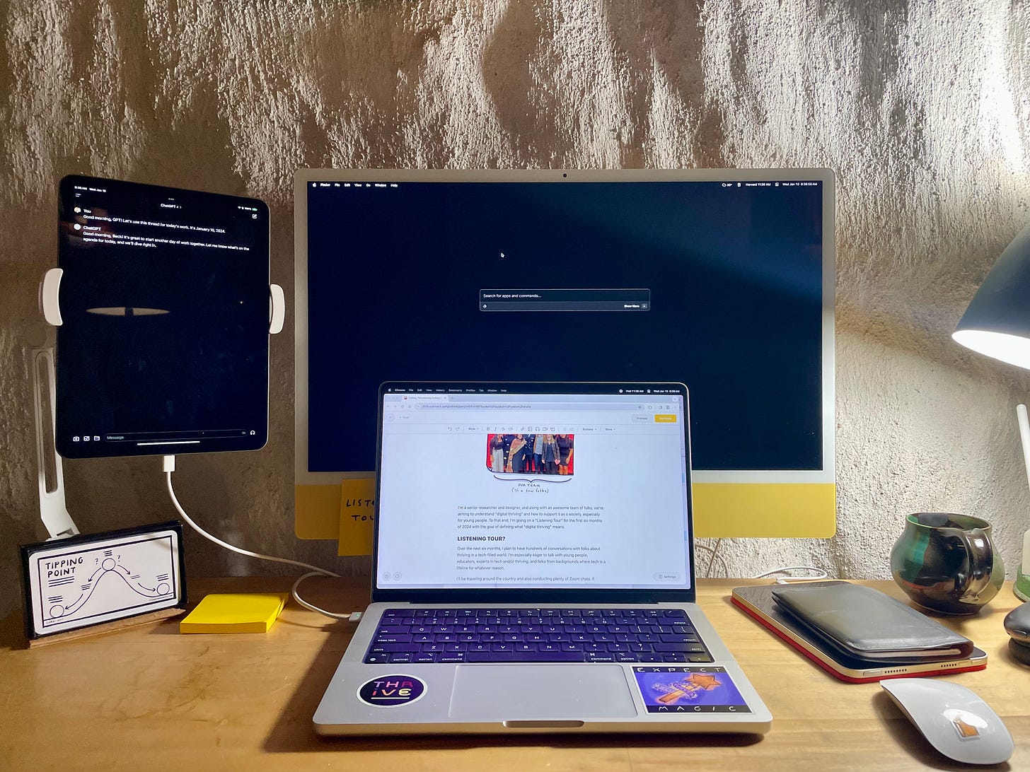 A well-organized workspace featuring a laptop open on a desk with a document on the screen, flanked by a tablet mounted on the left and a larger monitor on the right, both on standby mode. The desk has a notepad, a mug, and other personal items, and is illuminated by a desk lamp, creating a productive and personalized work environment.