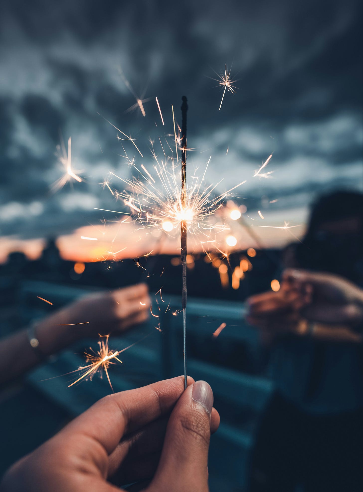 A hand holds a lit sparkler against a blurred out background. Slightly visible are three other people, each holding sparklers up, as if to toast each other. The distant background are light gray to dark blue clouds touching the top of a pink-orange sunset.