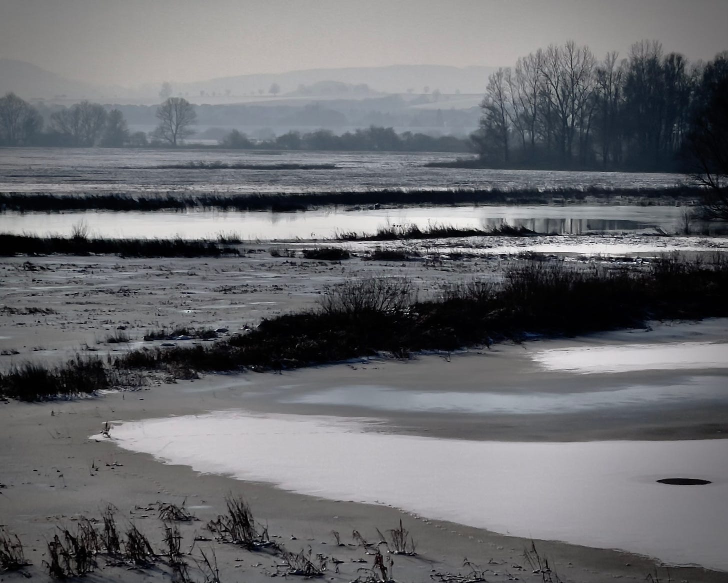 A serene winter scene of the flooded retention basin at Salzderhelden near Einbeck, Germany. Snow and ice cover the land, with reflective water cutting through barren fields and silhouetted trees standing against a hazy horizon. Photo by Jay Siegmann