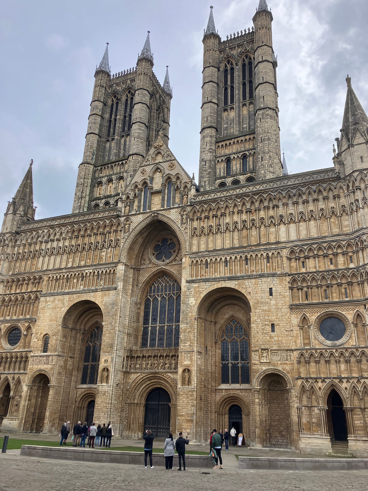 The frontage of the Lincoln Cathedral, showcasing Gothic architecture with two tall spires, a large rose window, and numerous ornate arches, with visitors at the entrance for scale.