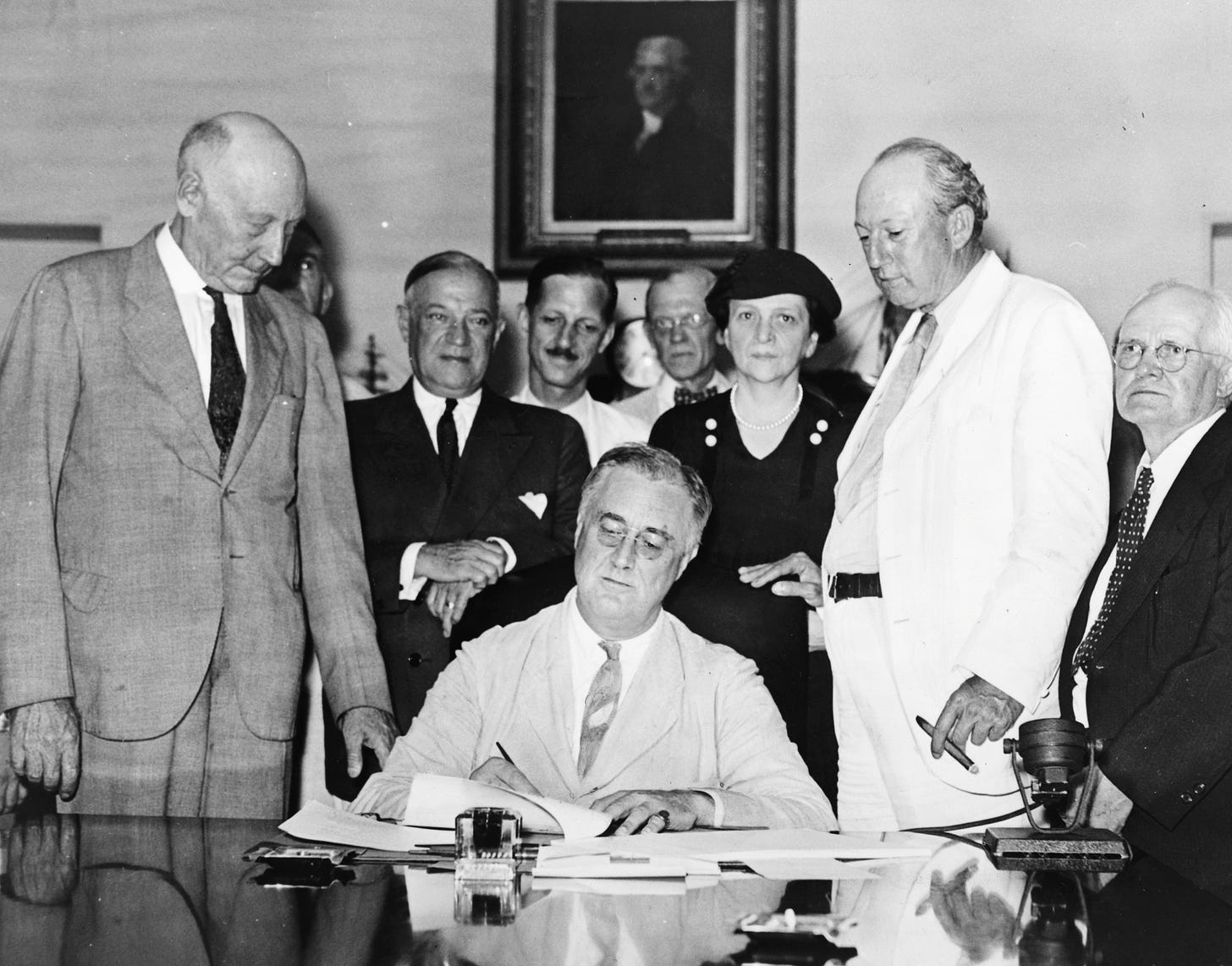 FDR seated, looking down at document he is signing while surrounded by several other people, mostly older white men. Portrait of Thomas Jefferson visible on wall behind them.