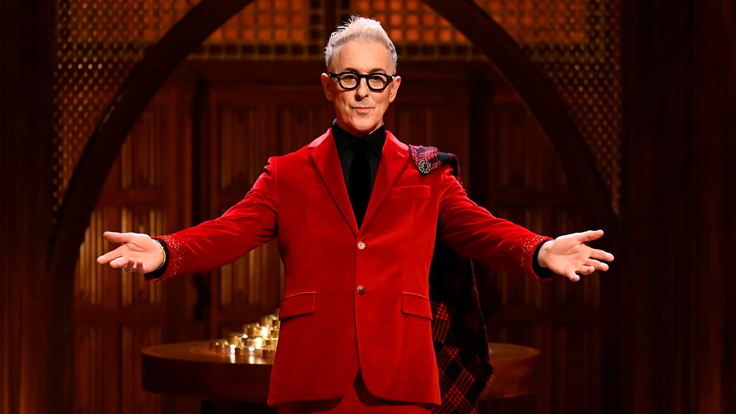 Alan Cumming in a red velvet suit with a plaid cape draped over his shoulder, standing with open arms in a wood-paneled room.