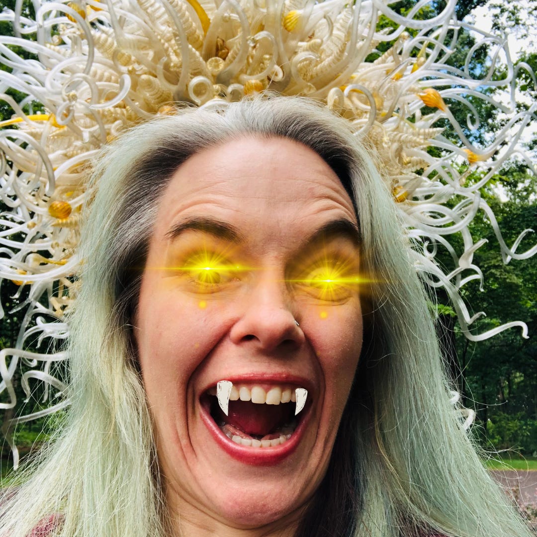 The smartass, green-haired author with fangs and golden flares for eyes in front of the corona of gold and white glass curlicues, horns and snakes.