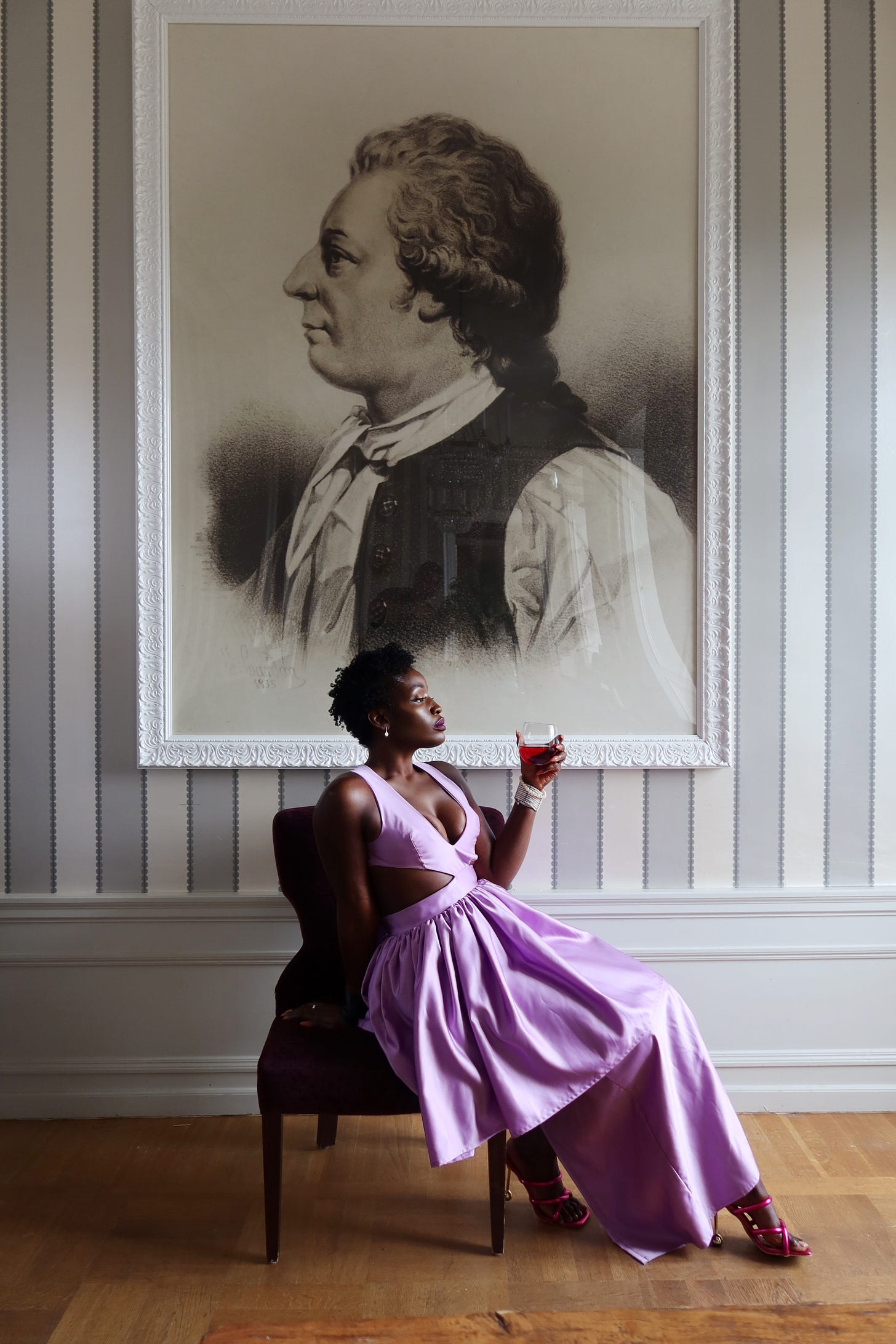 Lovette Jallow standing confidently in front of a historical portrait, glass in hand. Lovette Jallow standing confidently in front of a historical portrait, glass in hand.