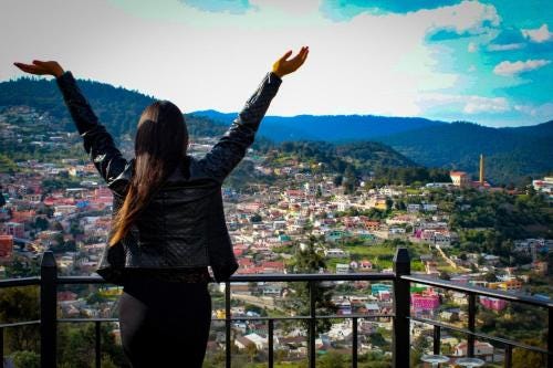 Woman standing with arms raised and hands spread out, facing green mountain view, dotted by red roofed homes. Standing on balcony of Hotel Santiurde, in Mineral del Monte, Hidalgo, Mexico.
