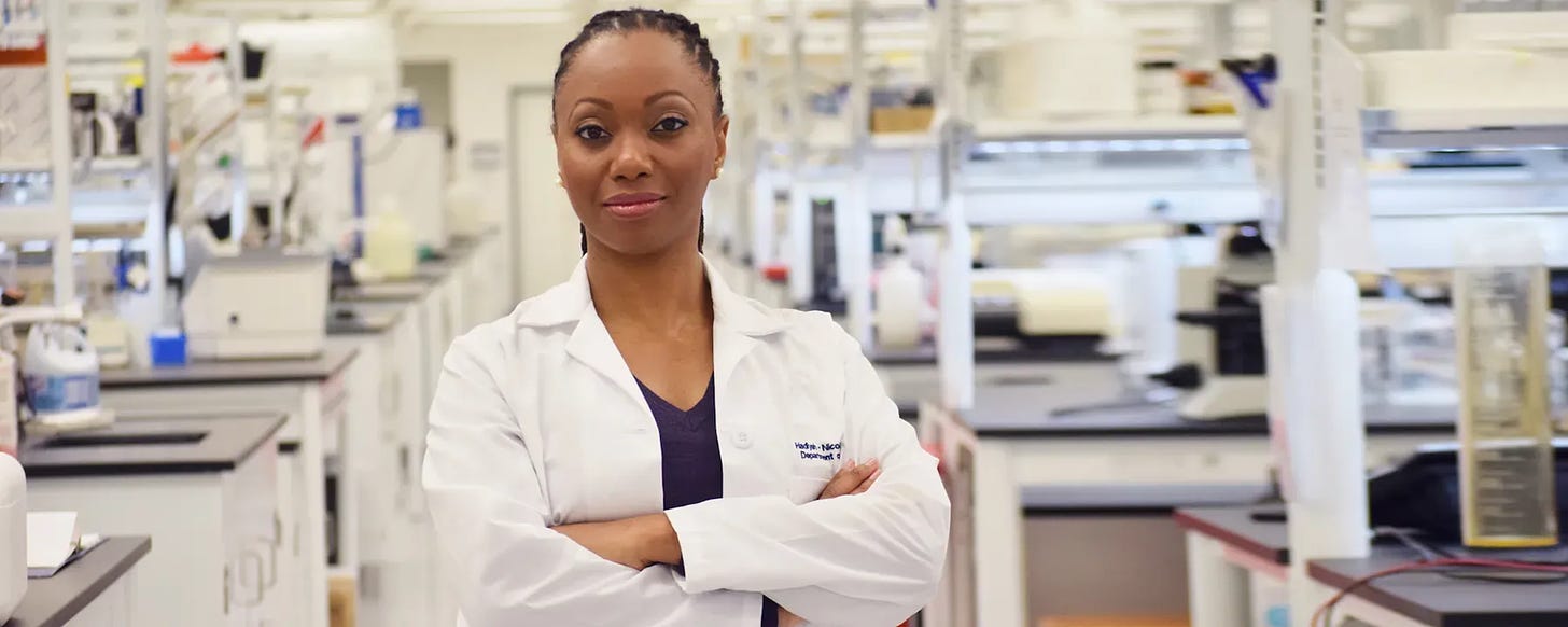 A Black woman stands in a lab. She's wearing a white lab coat with her arms crossed.