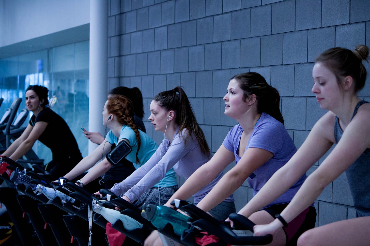 six women sit on six different spin bikes, all facing to the left. One woman has headphones in and is looking off to the side. 
