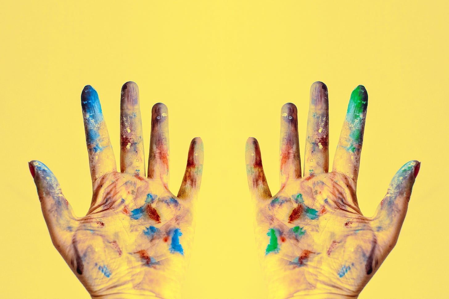 duas mãos, sujas de tinta de diversas cores no fundo amarelo