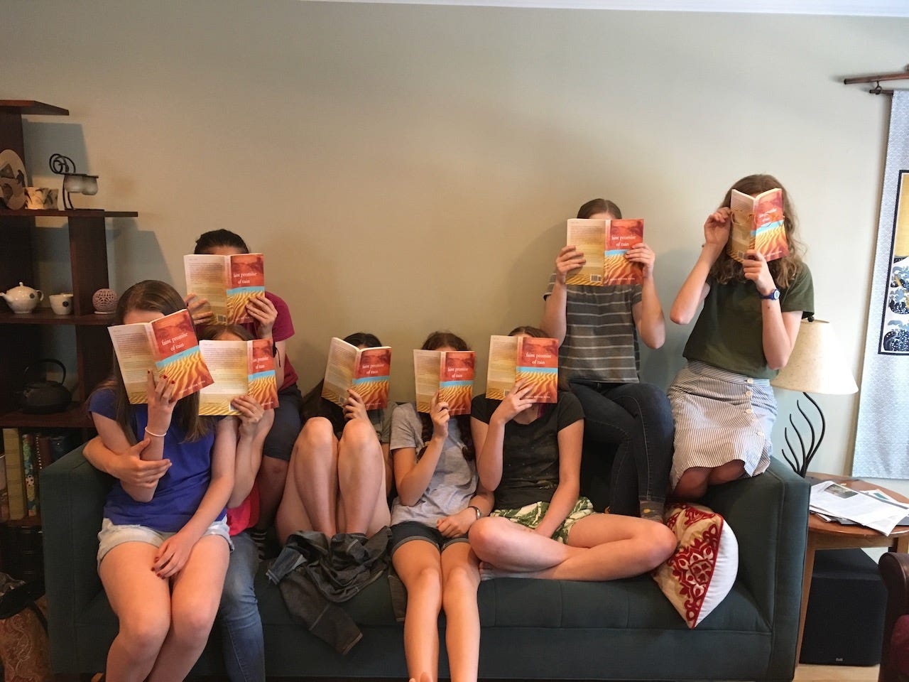 Seven teens sitting on a couch, faces covered by copies of the book Faint Promise of Rain