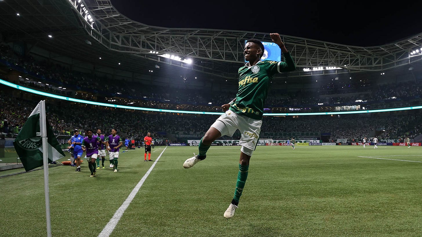 Estêvão, atacante do Palmeiras, comemora com braço erguido seu gol contra o Liverpool. Veste camisa e meias verdes e calção branco. É um jovem negro,com cabelos crespos. Ao fundo, se vê o telão do estádio.