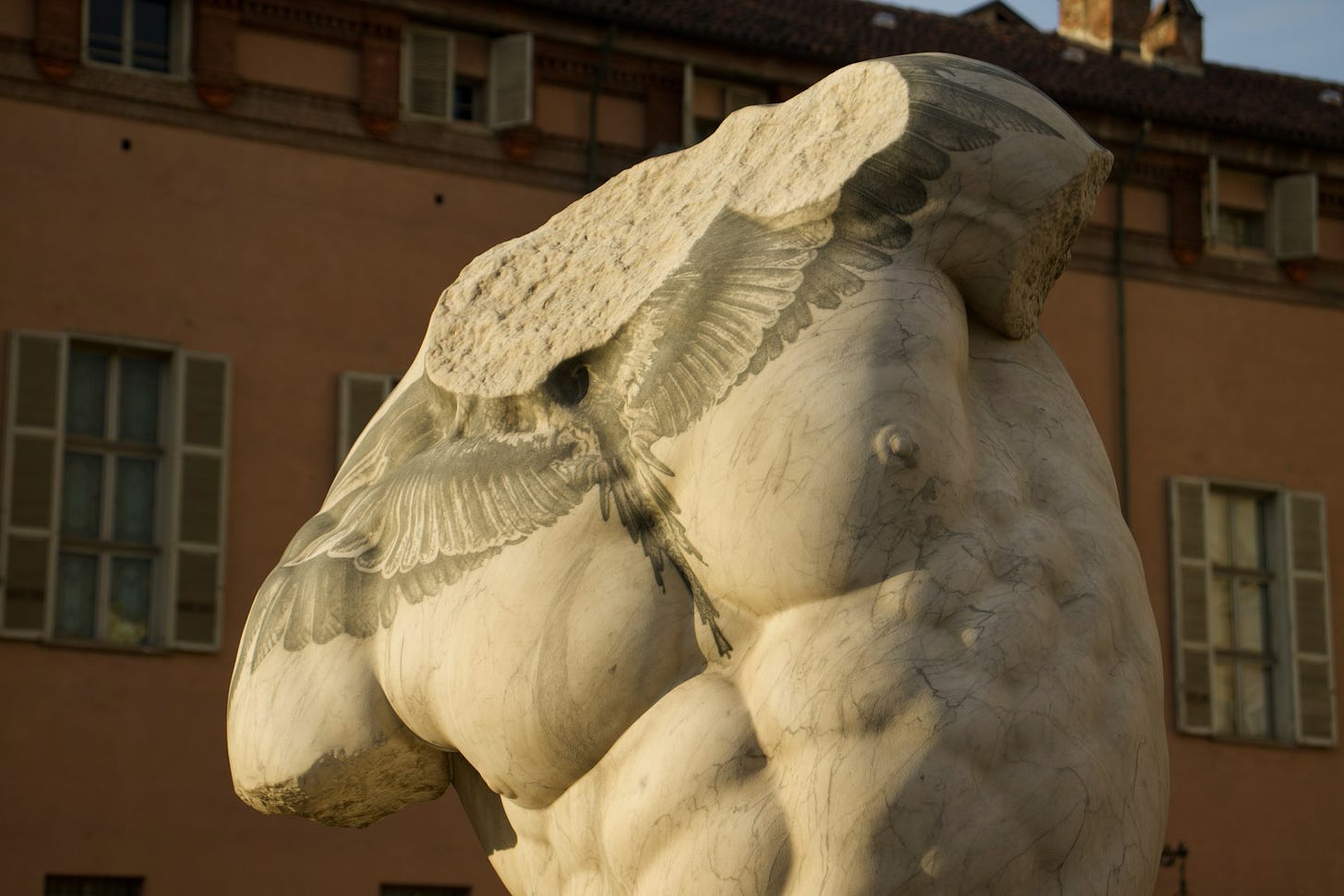 Escultura de mármore de um tórax masculino, sem a cabeça e os braços. O tórax, visto de frente e meio curbado para o lado esquerdo, tem uma tatuagem desbotada na parte superior do peitoral, de um par de asas compridas. Ao fundo, a fachada alaranjada de um prédio com janelas brancas.