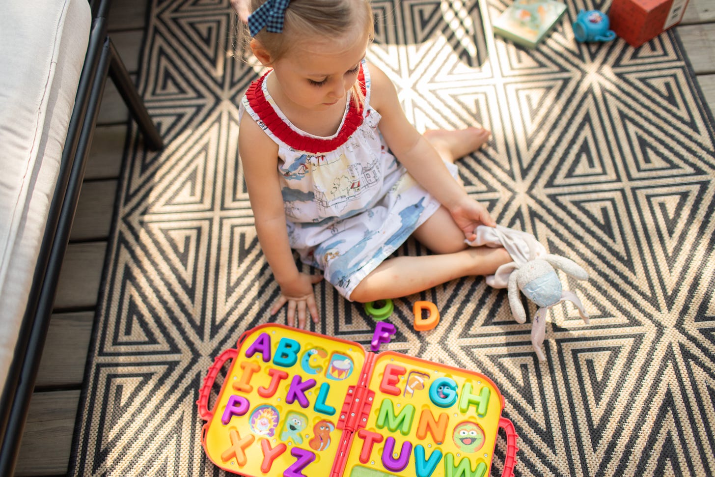 A child sits on an outdoor rug while holding a comfort item.