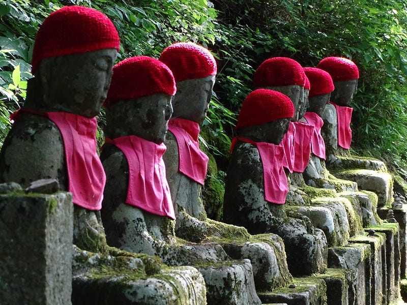 Image description: A row of statues, lichen-covered and worn, seated in peaceful mediation. Each wears a red knit hat and red bib..