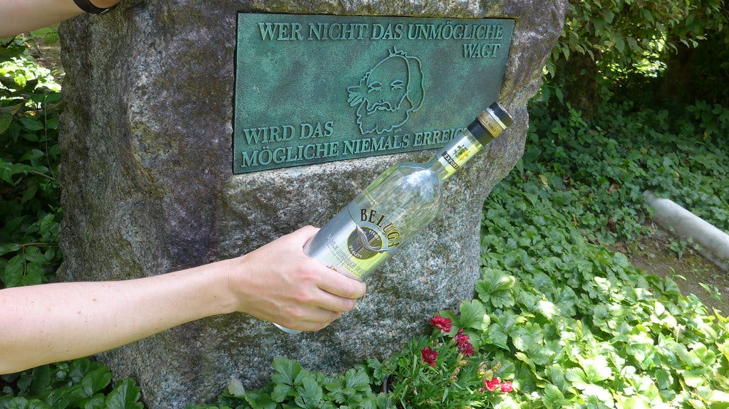 The grave stone of Bakunin with the engraving: Wer nicht das unmögliche wagt, wird das mögliche niemals erreichen. In Front there is a hand holding a Vodka bottle.