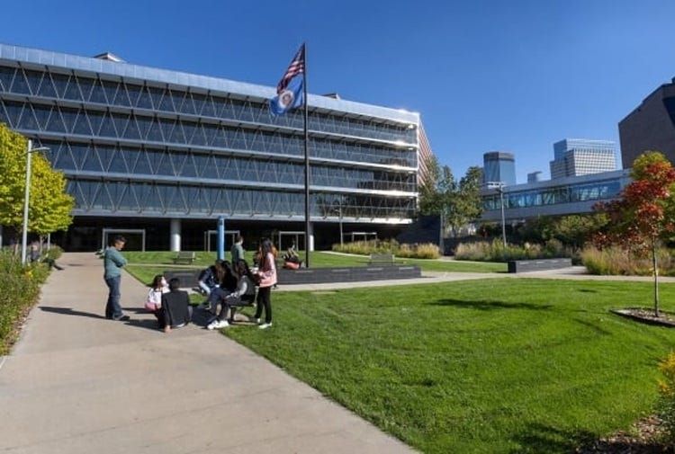 Students sitting on and around a bench have a conversation on a college campus with green lawns and academic buildings in the background.