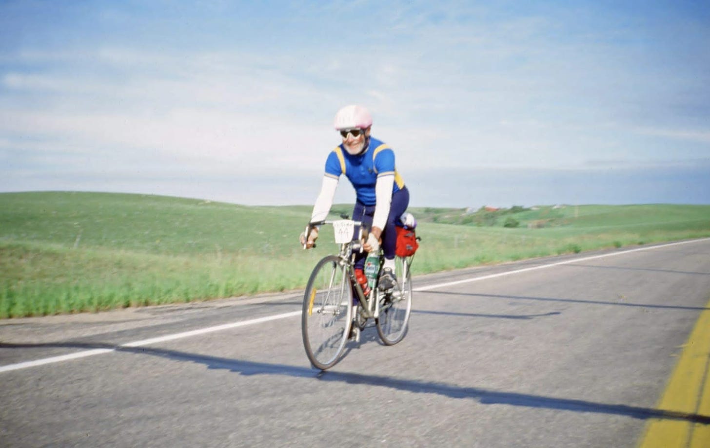 a single person wearing a blue shirt with white sleeves and navy pants and pink bicycle helmet rides a skinny-wheeled touring bicycle down an asphalt road with a yellow line visible on the right side of the frame and on the left a eternal field of green grassy and a white-and-light-blue striped sky behind