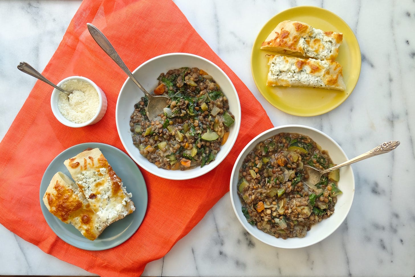 Two bowls of lentil stew served in white bowls with spoons sticking out, with slices of focaccia bread served on plates set out to the side, served on top of a marble countertop with an orange napkin