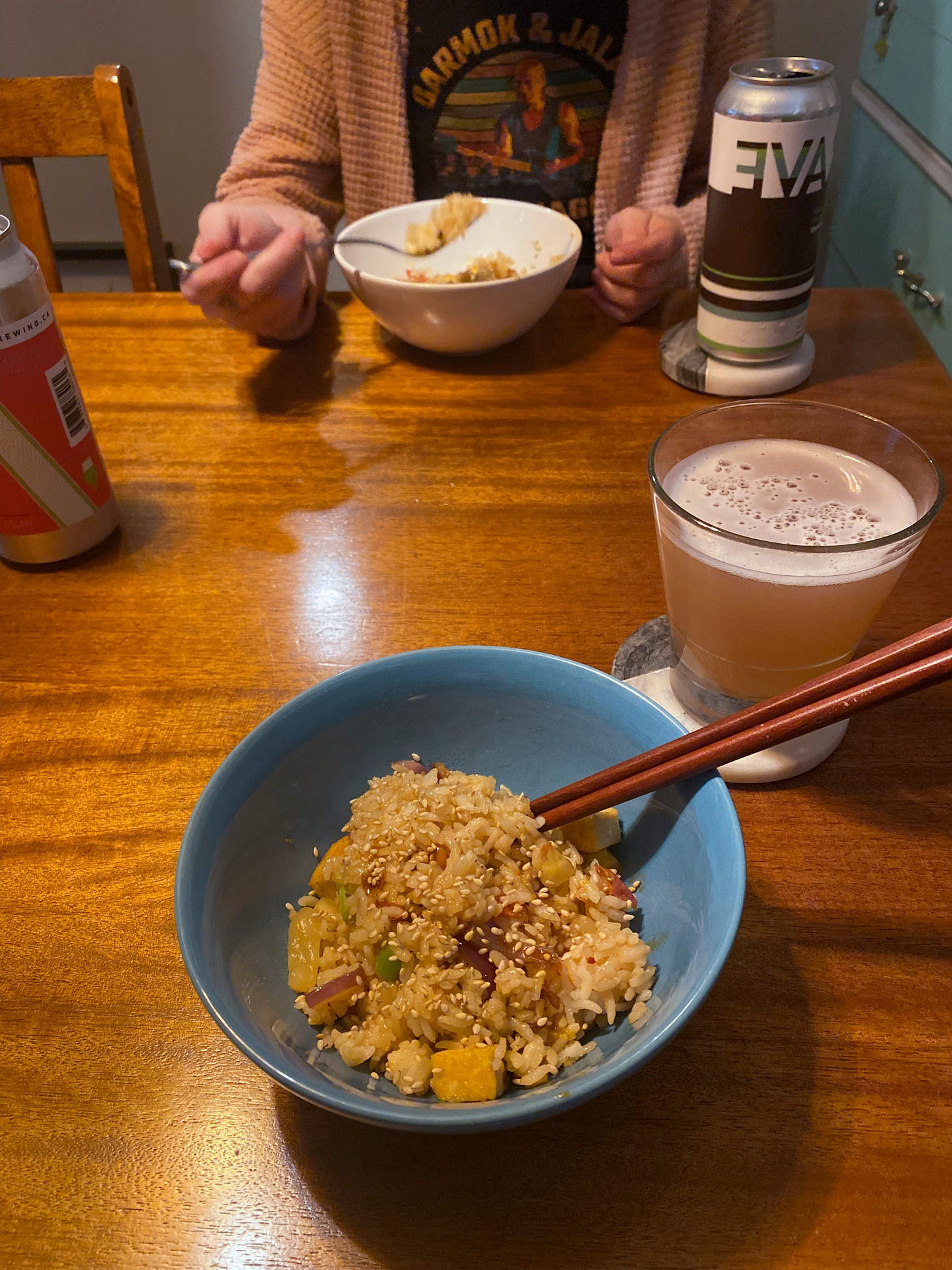 A blue bowl of fried rice topped with sesame seeds and chili oil, with wooden chopsticks at the side of the bowl. There's a glass of beer on a coaster beside it, and Steph sits in front of a white bowl of the same, with a can of beer on a coaster next to it. She's holding a fork over the bowl.