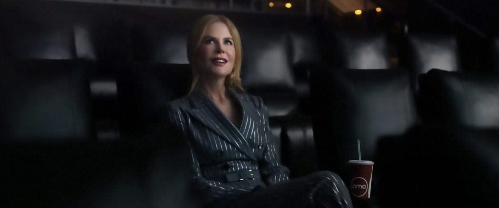 Nicole Kidman watching a movie in an empty theater whilst enjoying a fountain soda, wearing a black striped pantsuit.