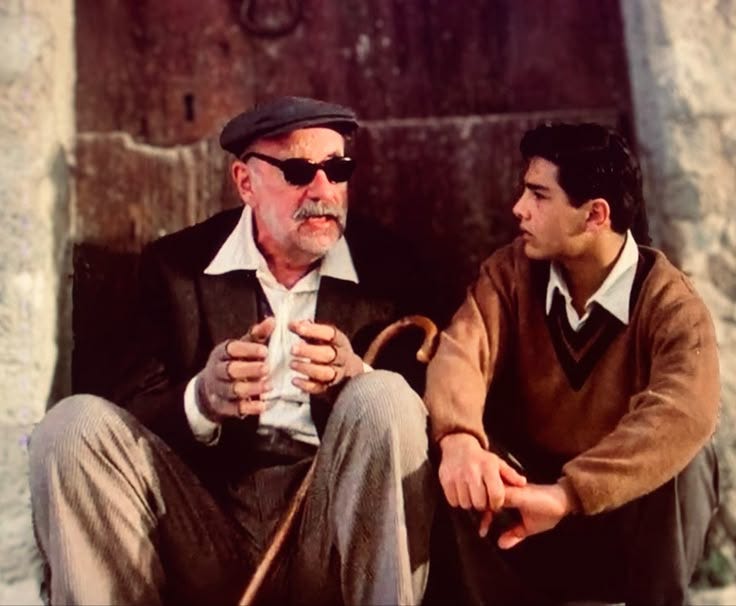 In a still from Cinema Paradiso, we an older man and a young man sitting on stone steps outside a house. The old man is talking, the young man is intently listening. In a still from Cinema Paradiso, we an older man and a young man sitting on stone steps outside a house. The old man is talking, the young man is intently listening.