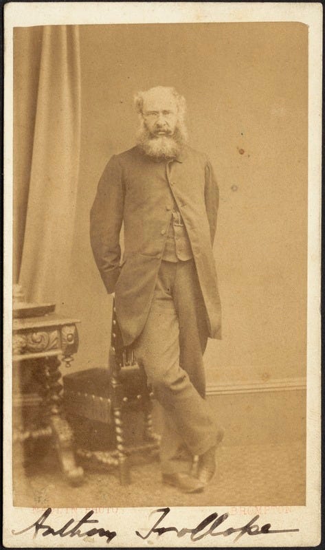 Photograph of Anthony Trollope leaning against a chair, his right leg crossed over the left. His name is written in ink at the base of the image.