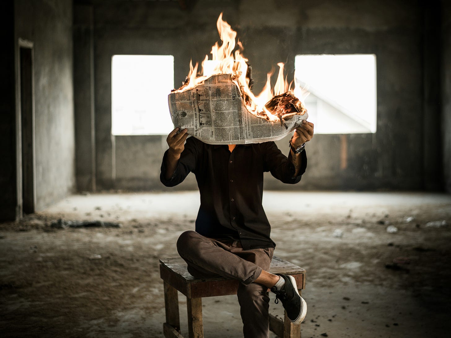 A person in a dark room, reading a newspaper. The newspaper is on fire.